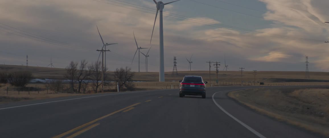 A car travels on a road surrounded by spinning wind turbines under a cloudy sky