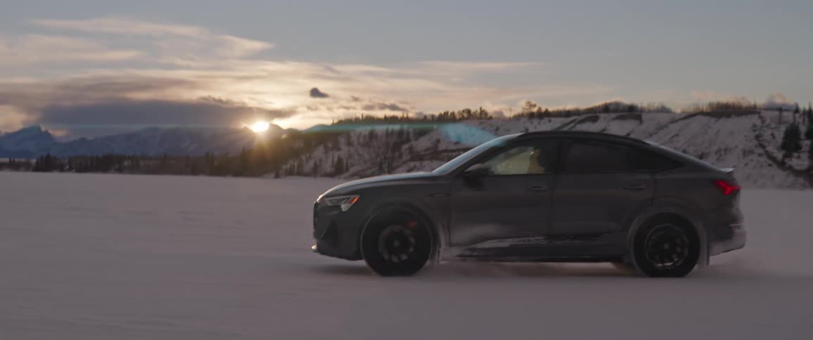A black car travels through the snow as the sun is setting