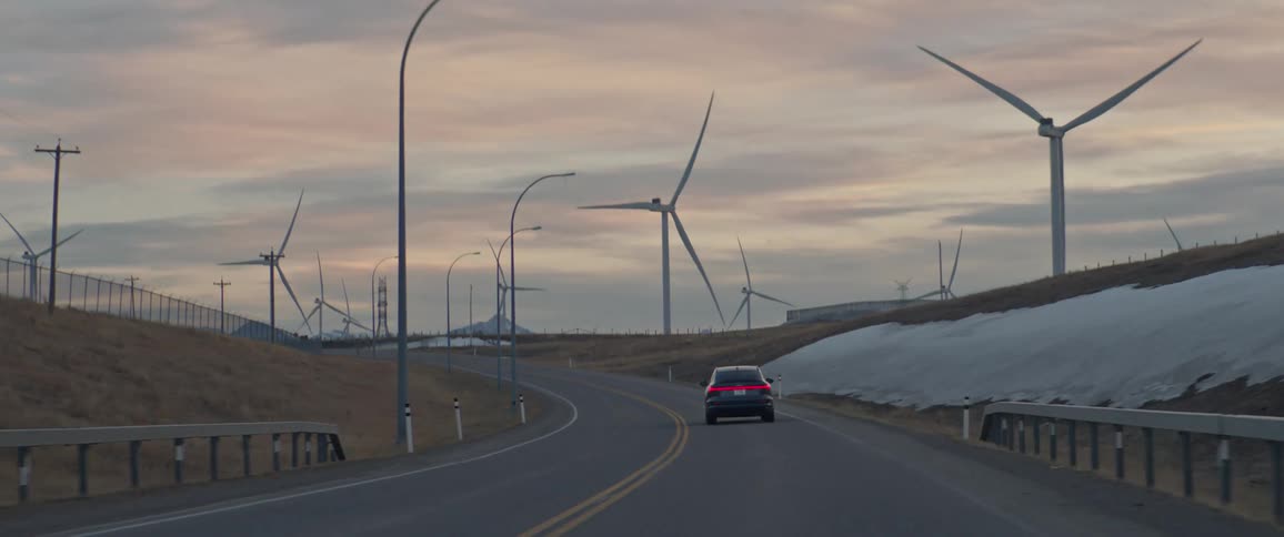 A car travels down a road towards a wind farm on a cloudy evening