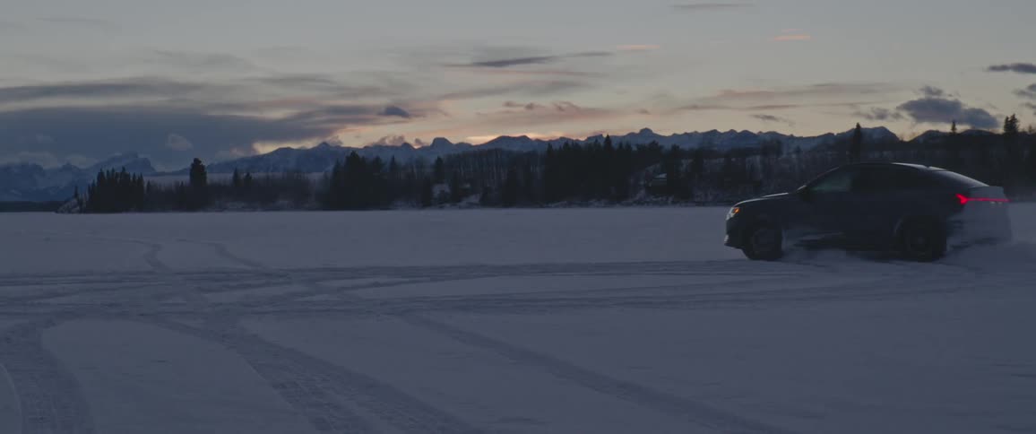 A vehicle drifting on a snowy field with a mountain range trees the sky and clouds in the background