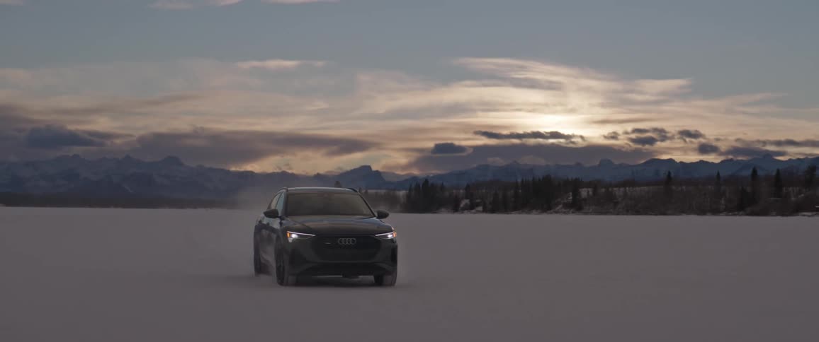 A black car travels through the snow as the sun is setting with mountains in the distance