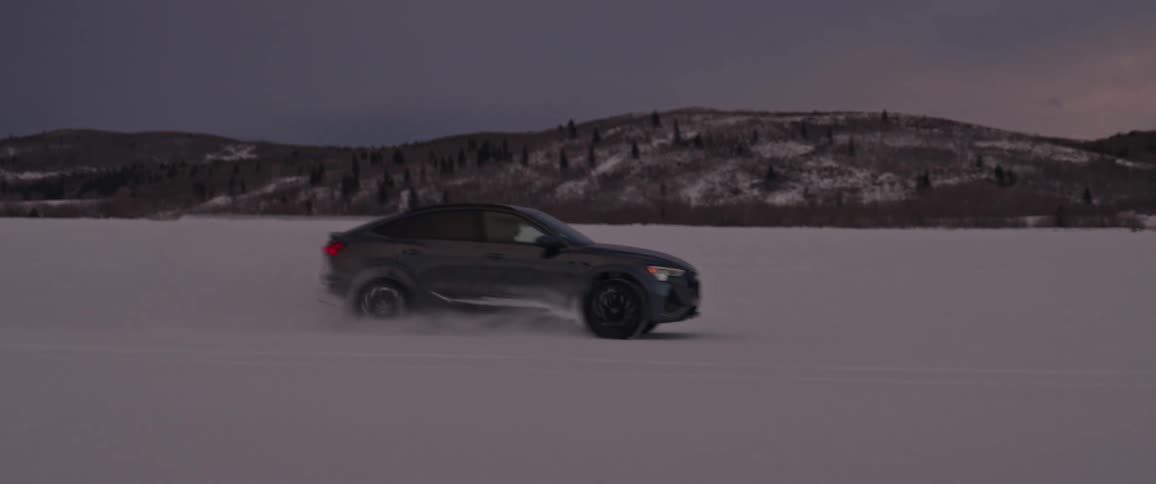 A black car travels through the snow with snow covered mountains in the distance