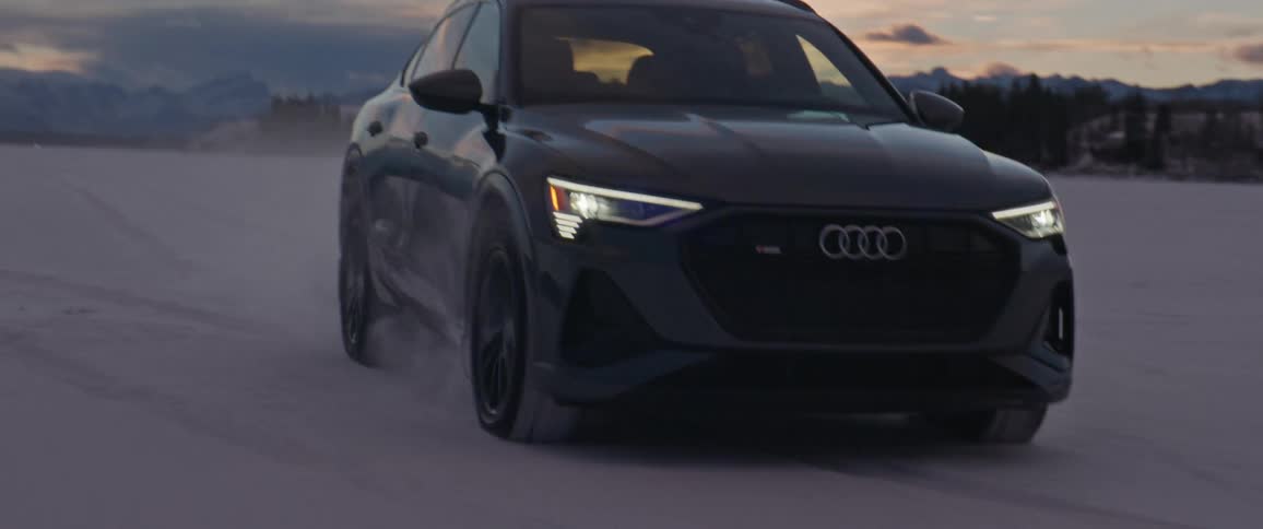 An audi vehicle drives along a large open snow covered area