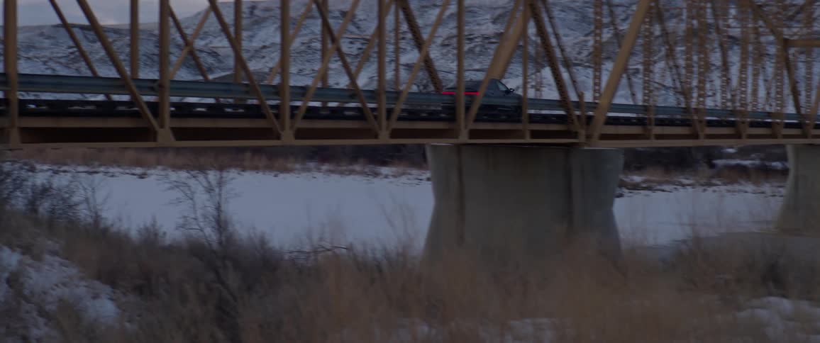 A car drives over a rural snow covered bridge beside a mountain