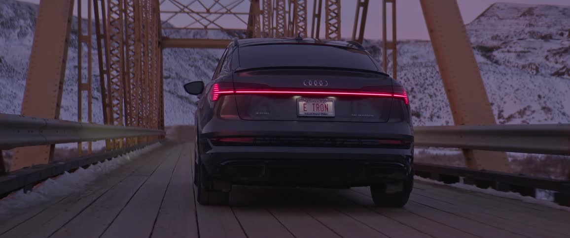 A black audi car is driving on a bridge with snowy mountains visible at a distance