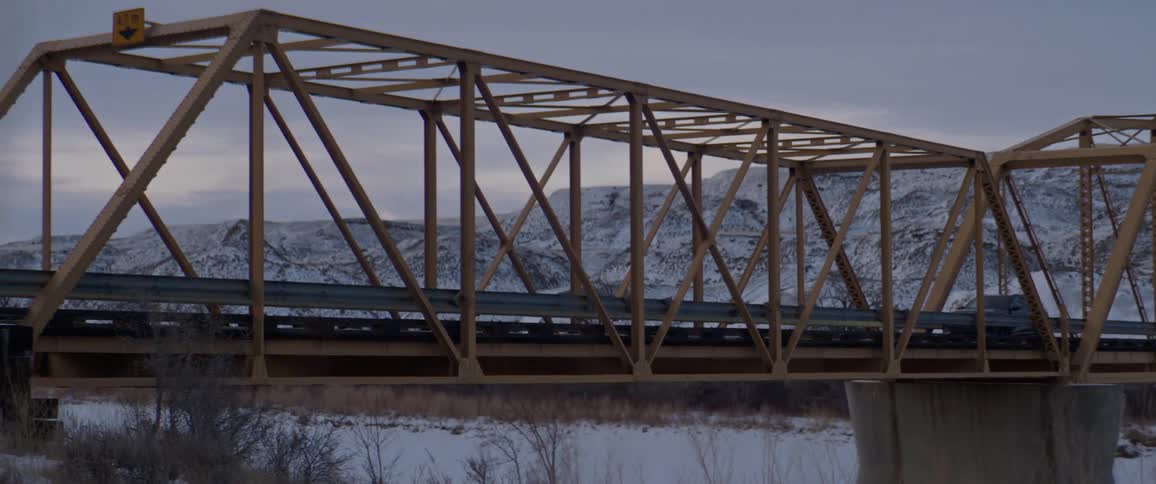 A vehicle travels across a bridge across a wintry landscape on a cloudy day