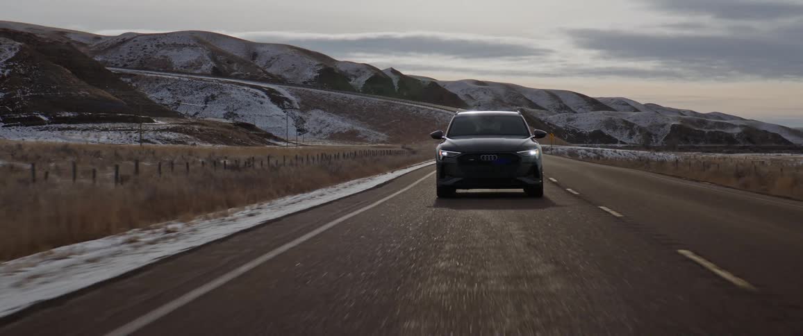 Dark grey audi car drives on highway with snowy hills in the background