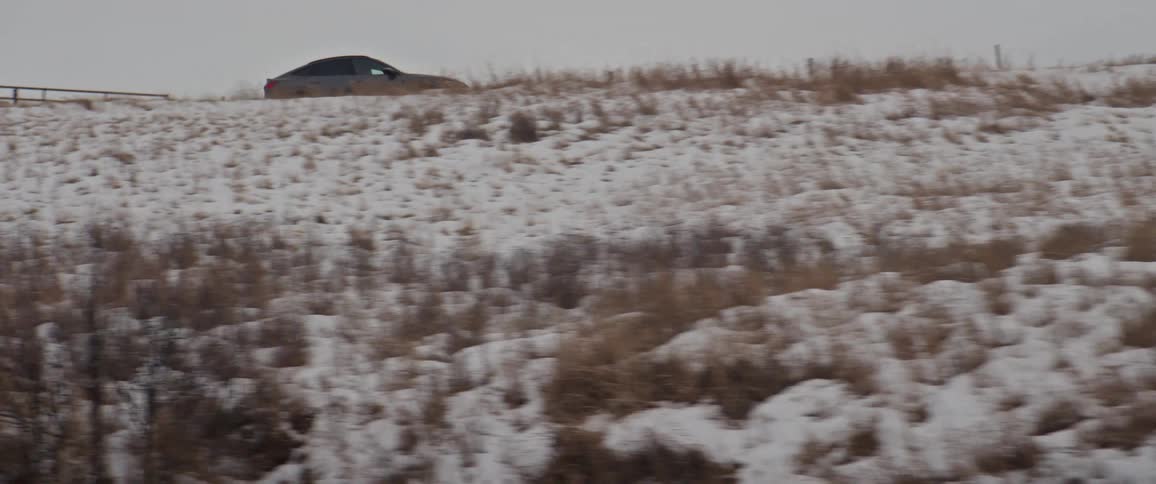 A vehicle driving with a fence and a snowy field with plants in the foreground with the sky and clouds in the background