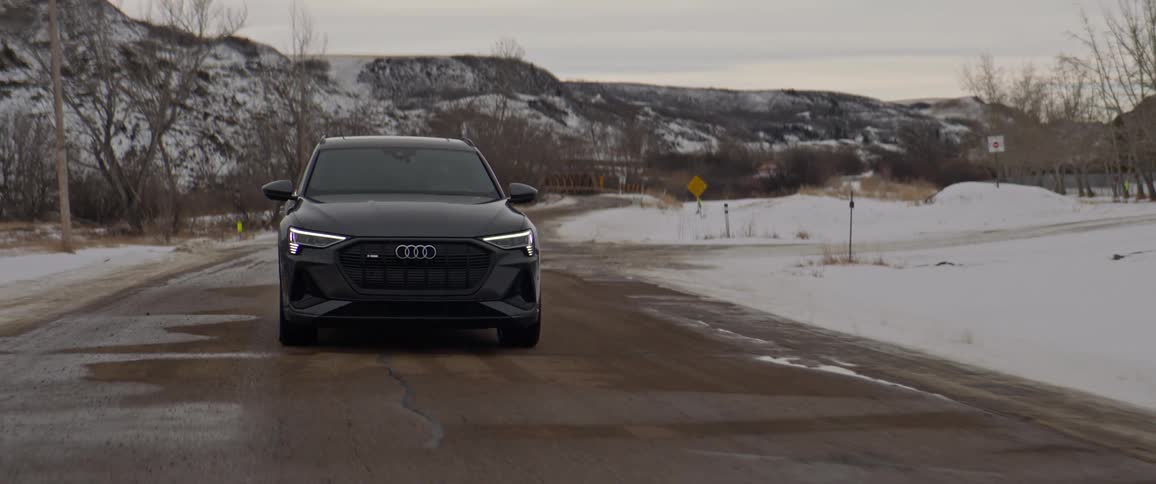 A black car travels on a road with snow and trees along the roadside and mountains in the distance