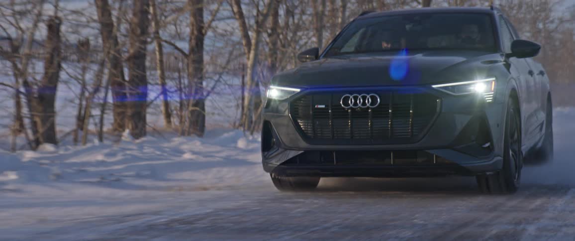 A black audi automobile travels on a snow covered road with trees along the roadside