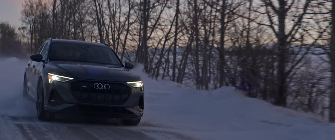 A black car travels on a snow covered road with headlights on and trees along the roadside