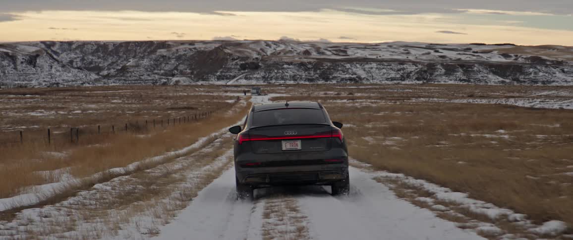 A black car travels on a snow covered road with snow covered mountains in the distance