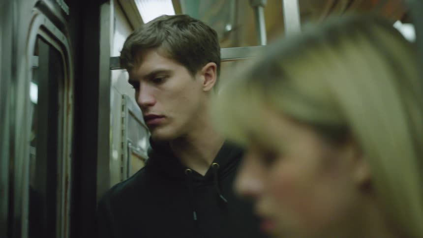 A young man is standing and looking outside as he travels in a train next to a woman