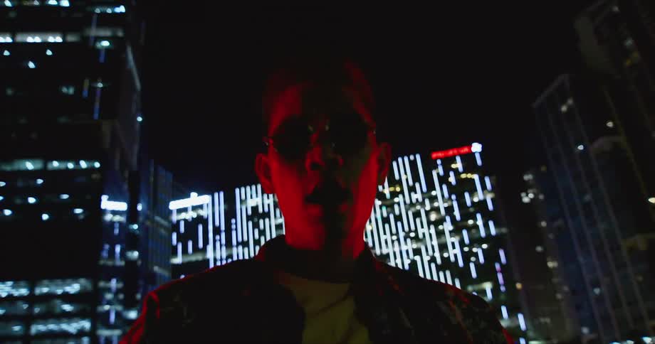 Young asian man wearing sunglasses standing in front of buildings in the city at night looks at the camera