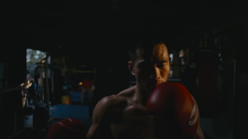 Shirtless man shadow boxes looking at the camera inside a dimly lit gym