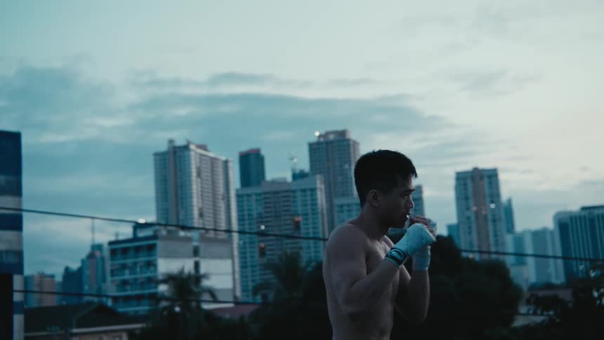 A shirtless male stands atop a building while practicing throwing punches at the air