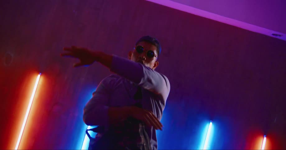 A man wears sunglasses indoors as he dances in a club with bright neon lights on the walls