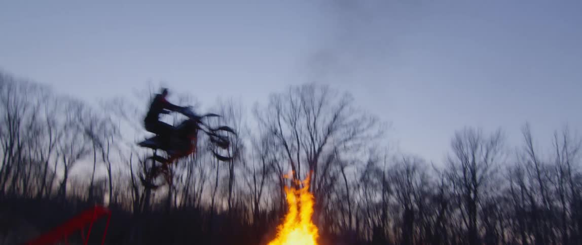 Person rides up ramp to jump over bonfire burning in field near woods full of leafless trees