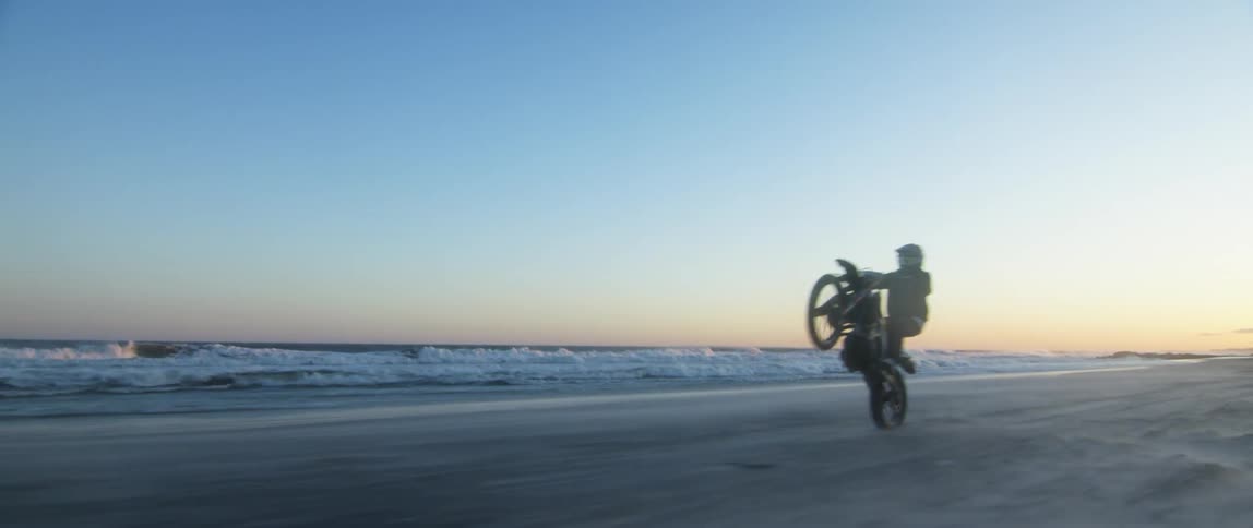 A motorcyclist rides a wheelie on a motorcycle as it is traveling on the beach near the surf
