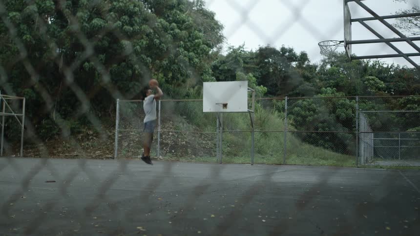 A man in a basketball court shooting a basketball with trees the sky and clouds in the background