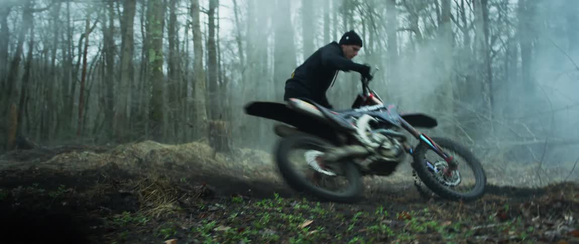 A man with a hat and hoodie revving a motorcycle in the mud in the forest with the sky and clouds in the background