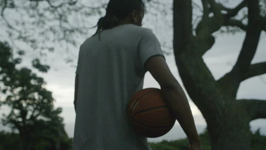 Man with braids and grey t shirt and shorts holds a basketball next to a tree