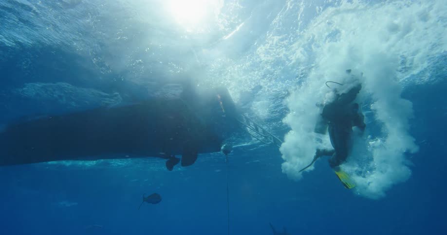 Scuba diver jumps into water from boat in cloud of bubbles as sun shines directly overhead and fish swim nearby