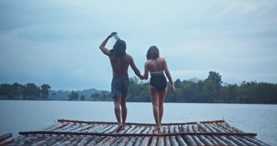 Couple wearing swim attire walk across wooden raft to waters edge holding hands on cloudy misty day