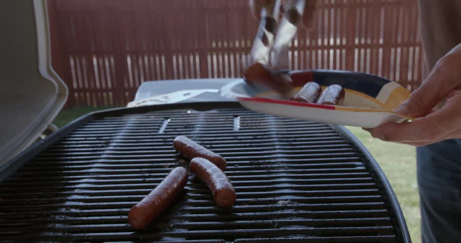 A man uses tongs to remove sausages from grill while cooking in his yard