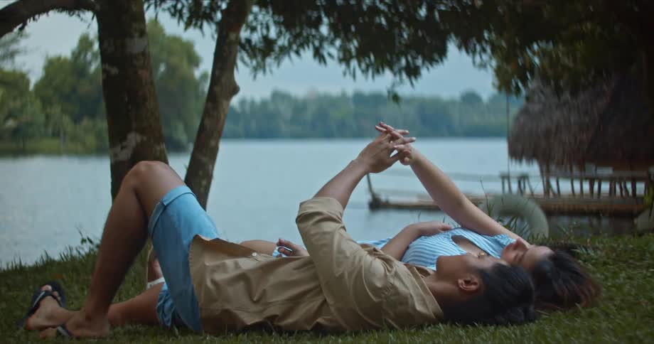 A couple is lying on grass lakeside and are holding hands