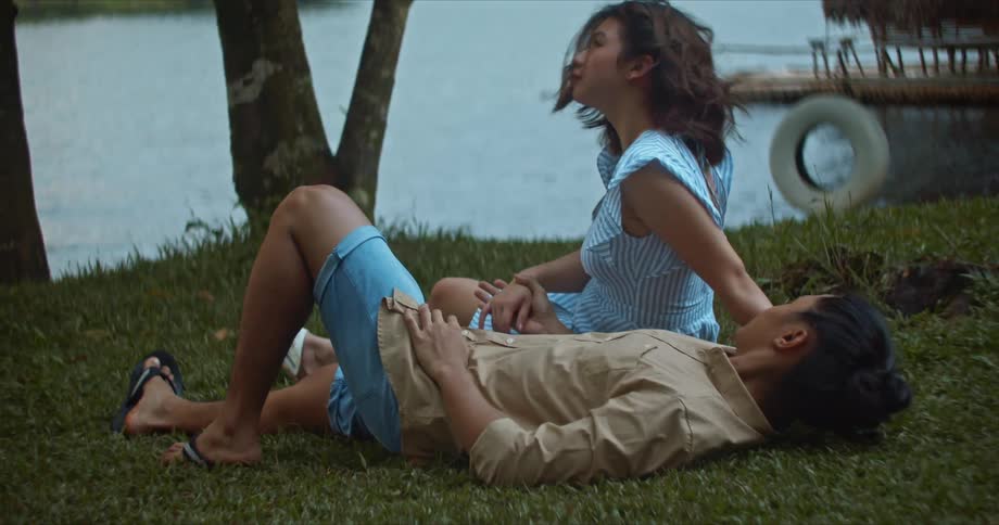 Couple rest on grassy riverbank holding hands with pier in background