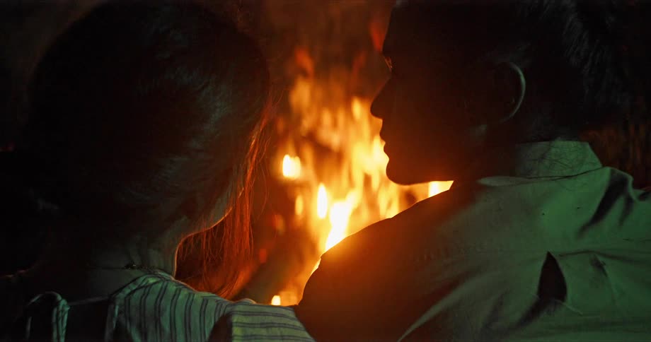 Couple sits side by side next to campfire and man stares at woman in the dark
