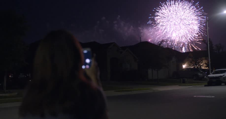 Woman stands in neighborhood street at night filming elaborate fireworks display with mobile phone
