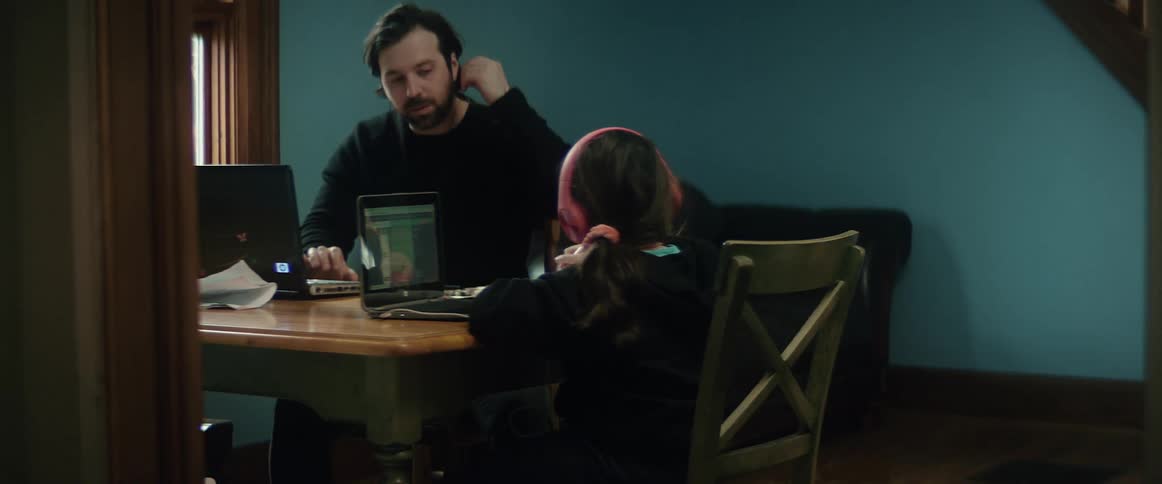 A man talks to a young girl as they sit together at a table using laptops during the day