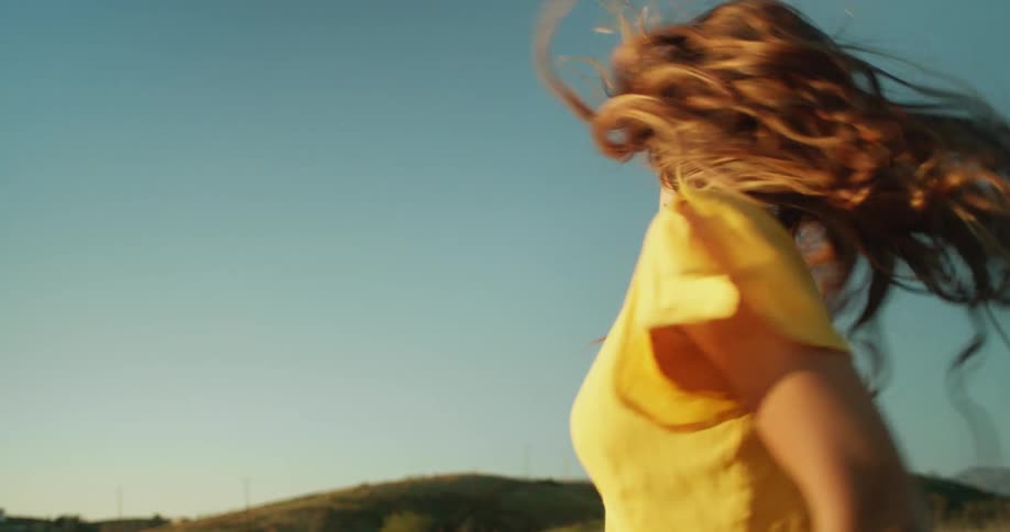 A woman with a yellow dress spinning and smiling with a landscape with hills the sky and clouds in the background