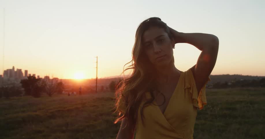 Woman wearing a yellow dress standing on a field looks at the camera at sunset