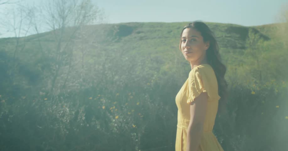 Woman wearing a yellow dress walking near a hill looks at the camera