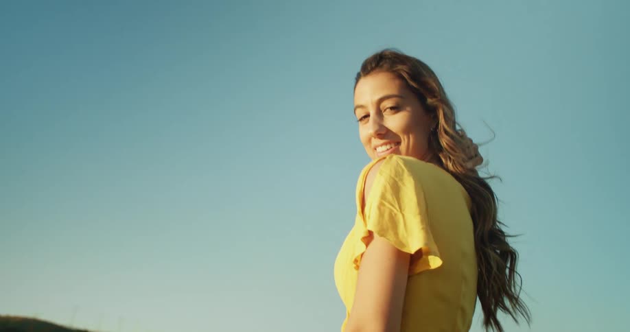 Woman with brown wavy hair and yellow dress poses for the camera with blue cloudless sky in the background