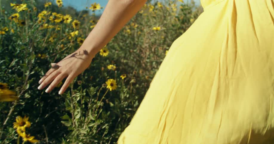 A woman with a yellow dress touching flowers and plants with the sky and clouds in the background