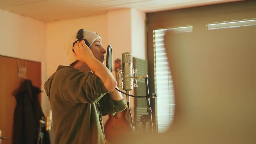 Male singer with white beanie and green hoodie wears headphones and sings a song in the studio