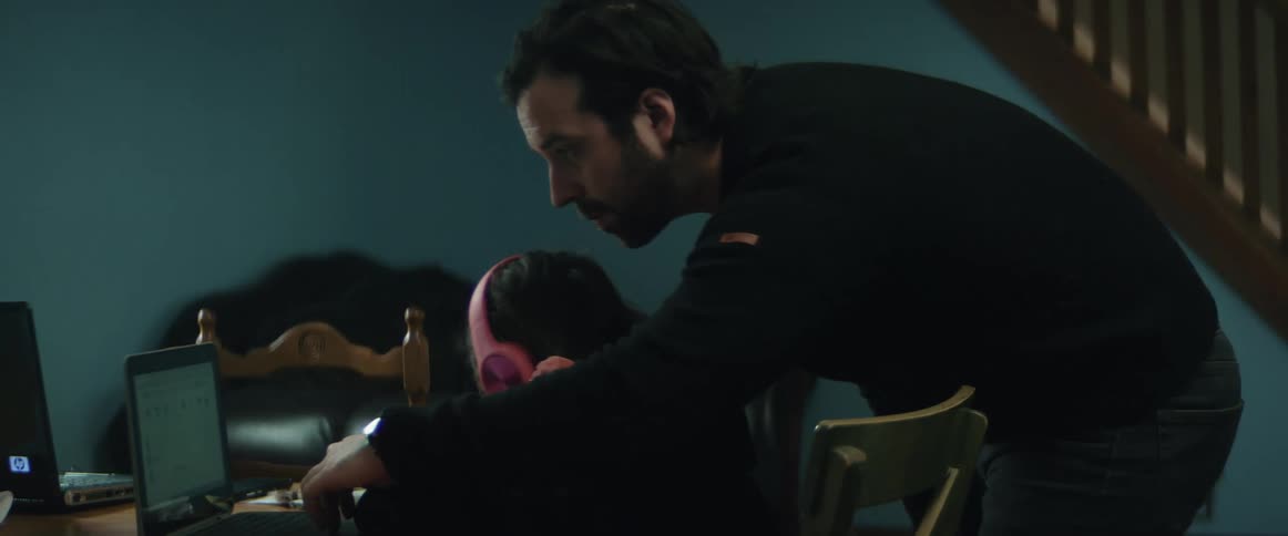 Father with beard and black sweater helps daughter wearing pink headphones study and sits down at table