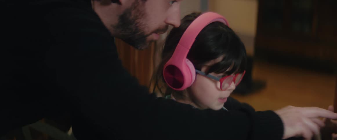 Father with a beard and a black sweater helps daughter wearing pink headphones use laptop at home