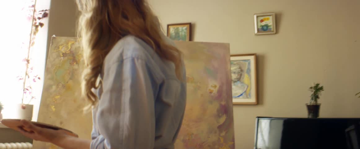 A woman uses a palette and brush to paint a canvas on an easel in a living room