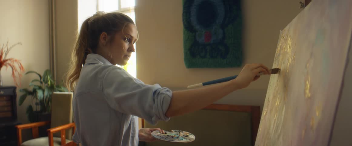 A woman tilts her head and examines her canvas as she paints