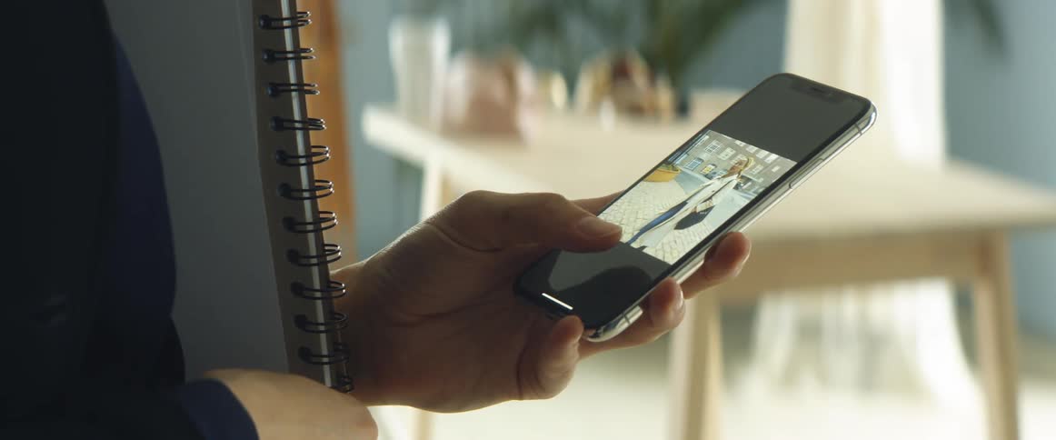 A woman scrolls through photos on her smartphone while standing with a notepad under her arm