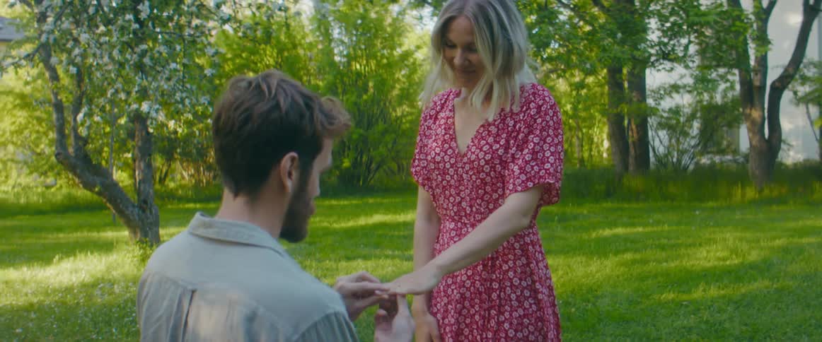 A man kneels to propose to a woman and places a ring on her finger before standing and sharing a kiss