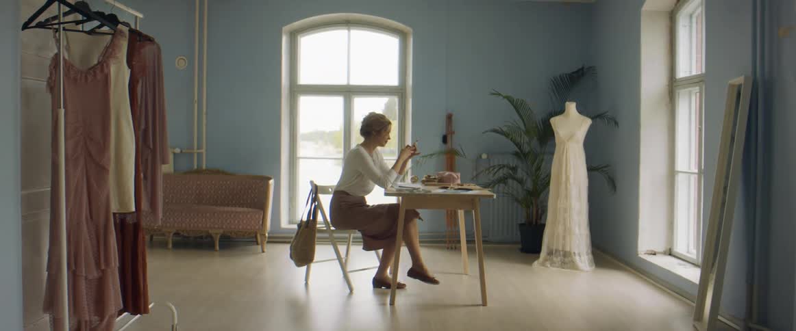 A woman in a room sitting on a chair at a table with a wall and window in the background