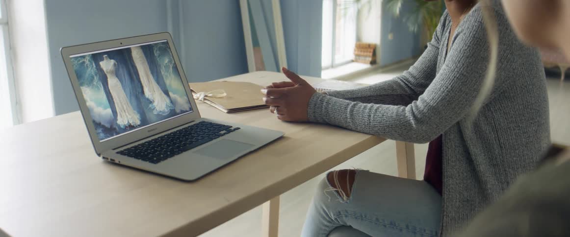 Two women sit at a table in a design studio looking at a laptop with a wedding gown displayed