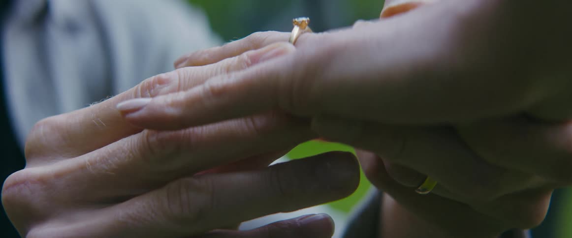 A man placing a ring on a womans finger with trees and grass in the background