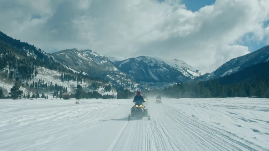 People driving snow mobiles across a snowy field with mountains in the background